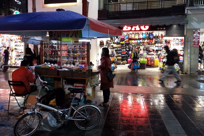 A rainy night in Taipei, Taiwan, where a small street vendor sells shoes and a cyclist waits under a blue umbrella.